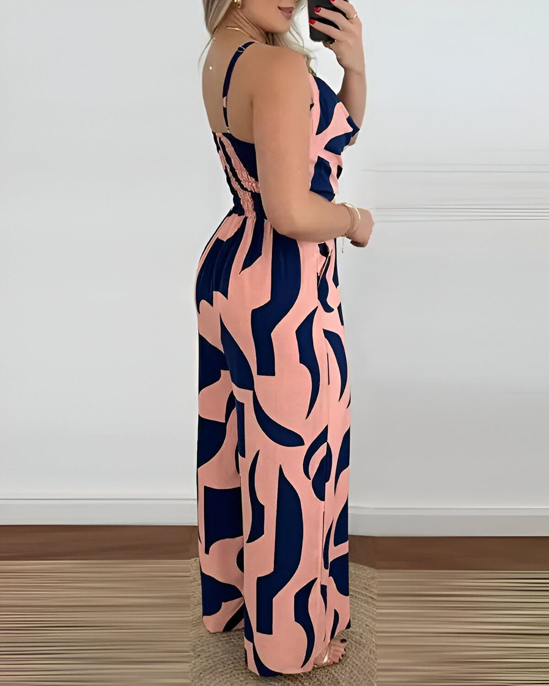 Une femme porte une combinaison pantalon rose et bleu marine. La combinaison a un haut court et un pantalon large. Le haut a des bretelles fines et un décolleté en V. Le pantalon est taille haute et a des jambes larges. La femme est debout, de côté, et tient son téléphone portable à la main.