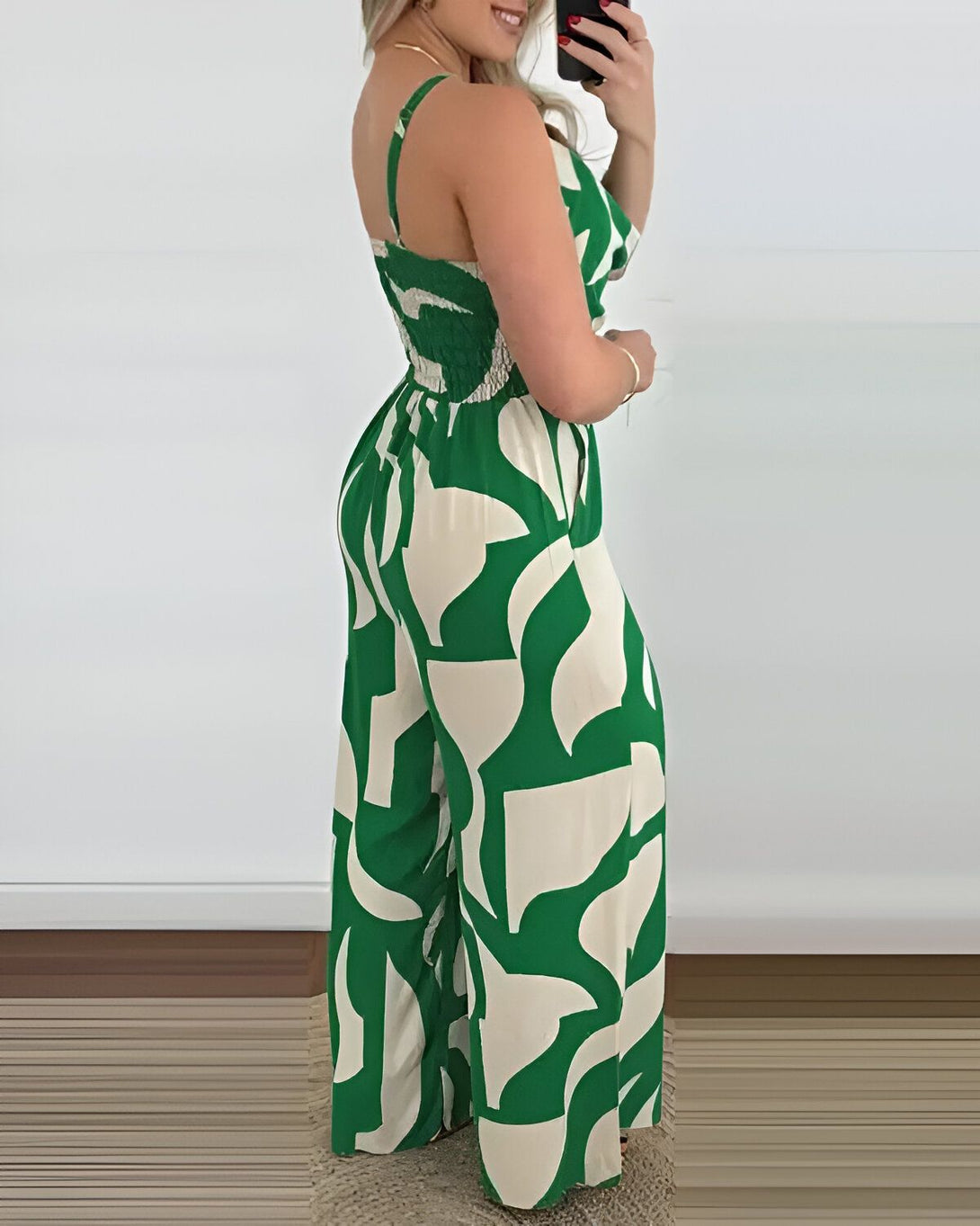 Une femme porte une combinaison pantalon verte et blanche à motifs géométriques. La combinaison est cintrée à la taille et possède des jambes larges. La femme est vue de dos et porte des talons hauts blancs.