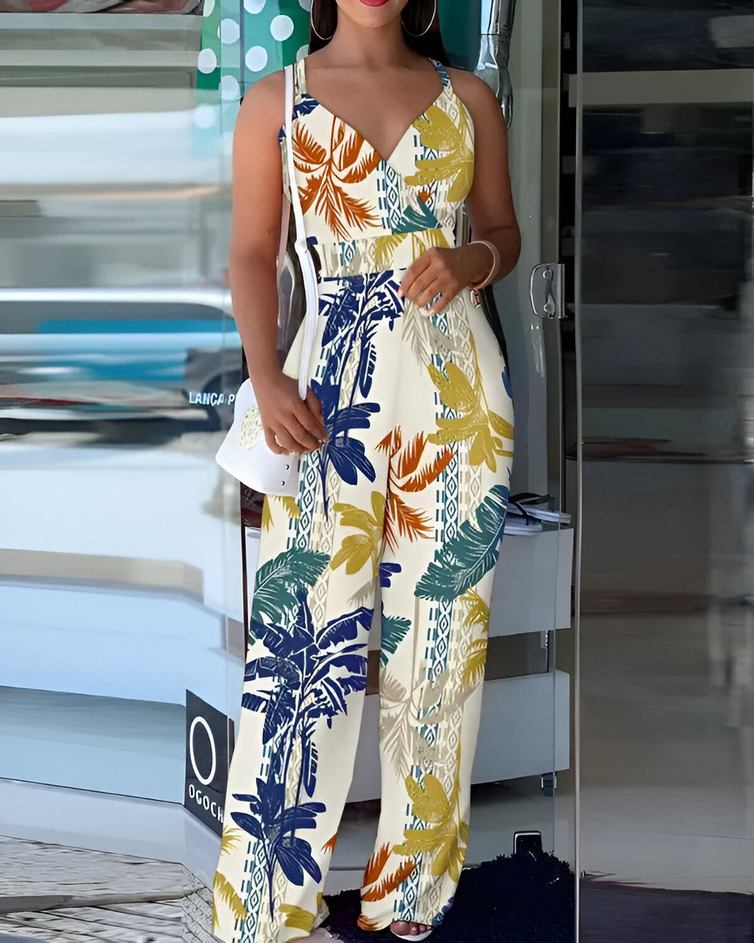 Une femme porte une combinaison pantalon ample et colorée à motifs de feuilles tropicales. Elle est dos à la caméra et porte un sac à main blanc.