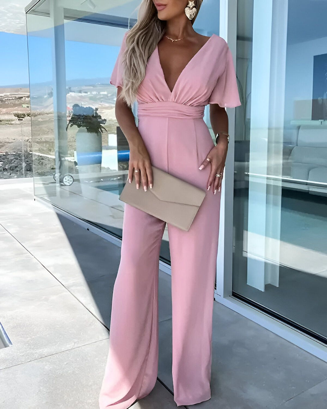 Une femme porte une combinaison pantalon rose avec un décolleté en V et des manches courtes. Elle porte également des talons hauts beige et une pochette beige.