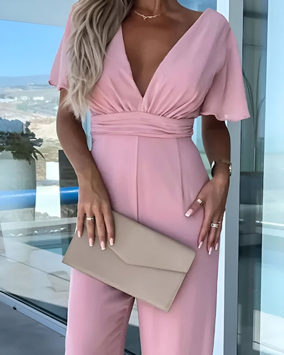 Une femme porte une combinaison rose avec un décolleté en V et des manches courtes. Elle porte également une pochette beige à la main.