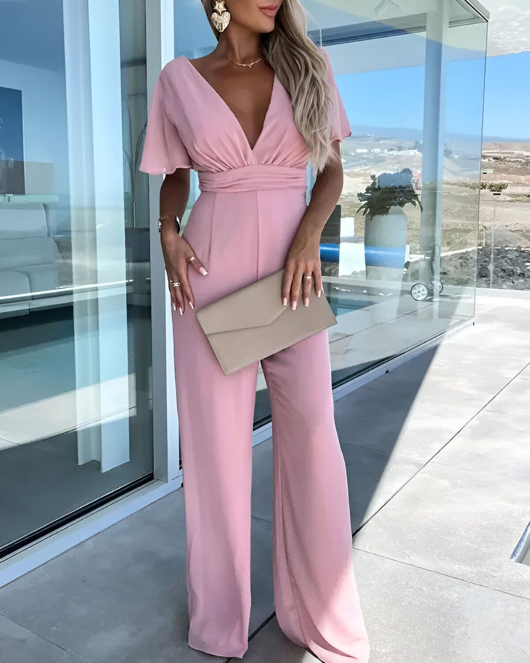 Une femme porte une combinaison pantalon rose. Elle est décolletée et a des manches courtes. Elle porte des talons hauts et un collier. Elle a une pochette à la main.