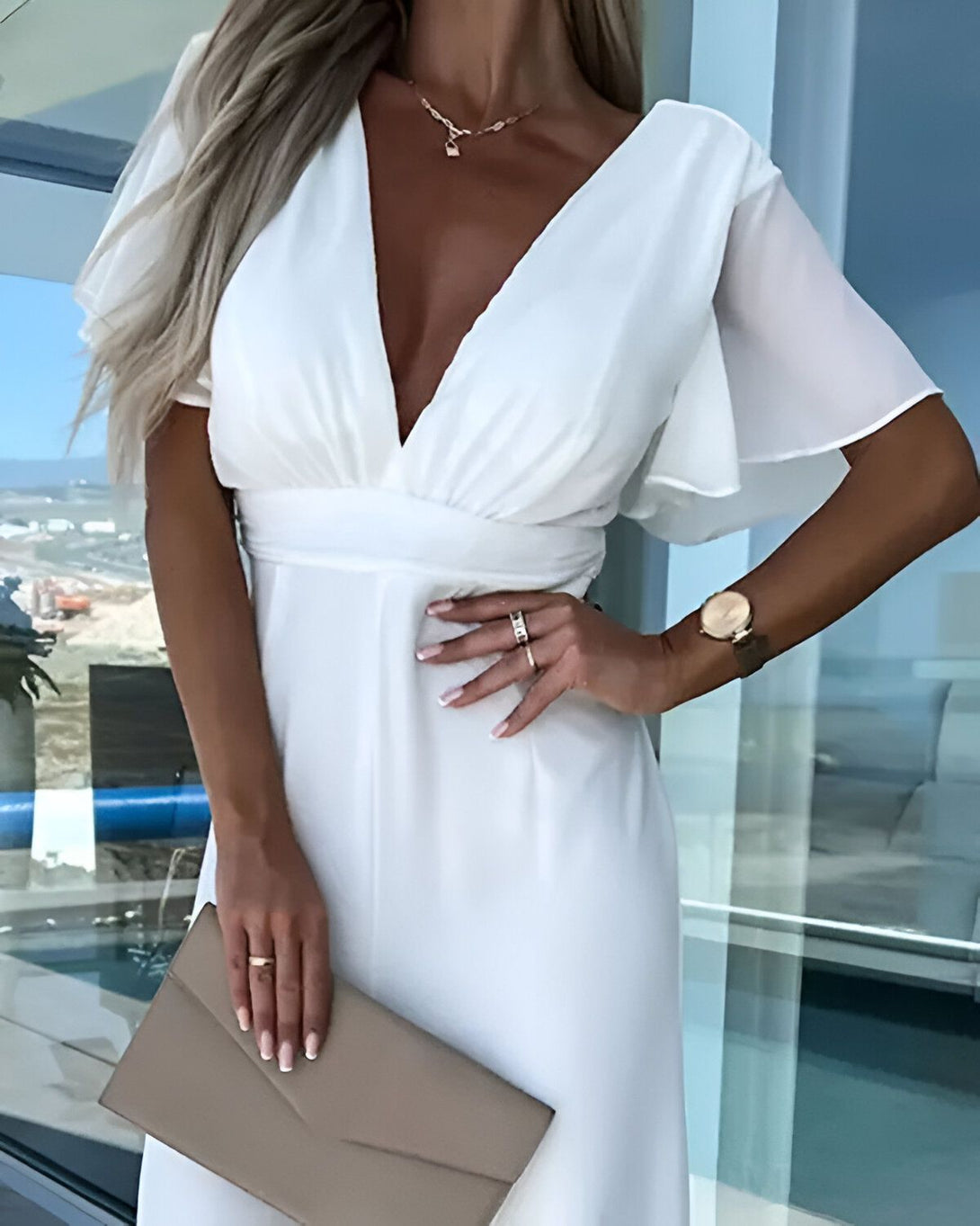 Une femme porte une combinaison blanche avec un décolleté plongeant et des manches courtes. Elle porte également une montre marron et un collier avec un pendentif en forme de cadenas. Elle tient un sac à main beige à la main.