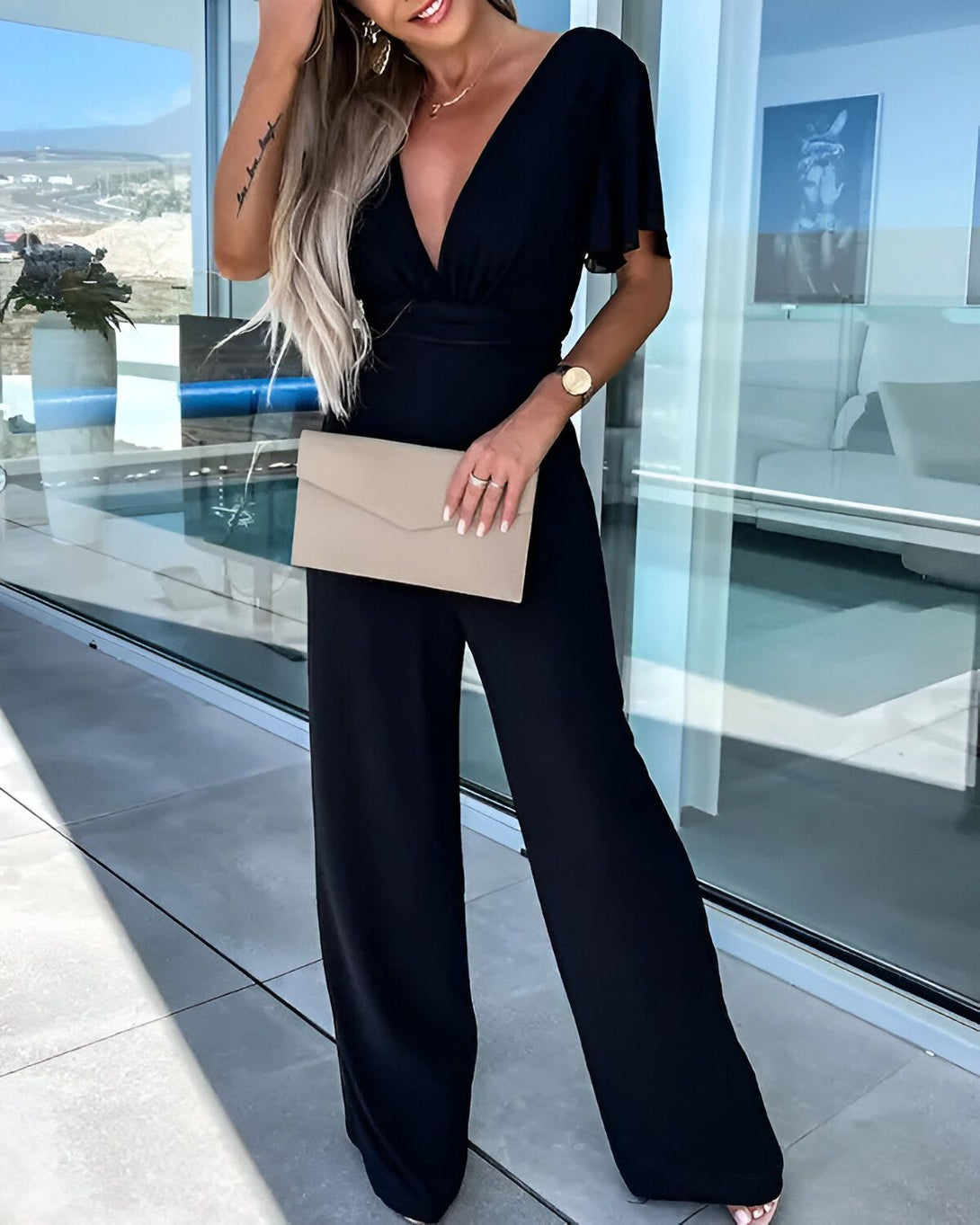 Une femme porte une combinaison pantalon noire décolleté en V et des manches courtes bouffantes. Elle porte également des sandales à talons beige et une montre. Elle a une pochette beige à la main.