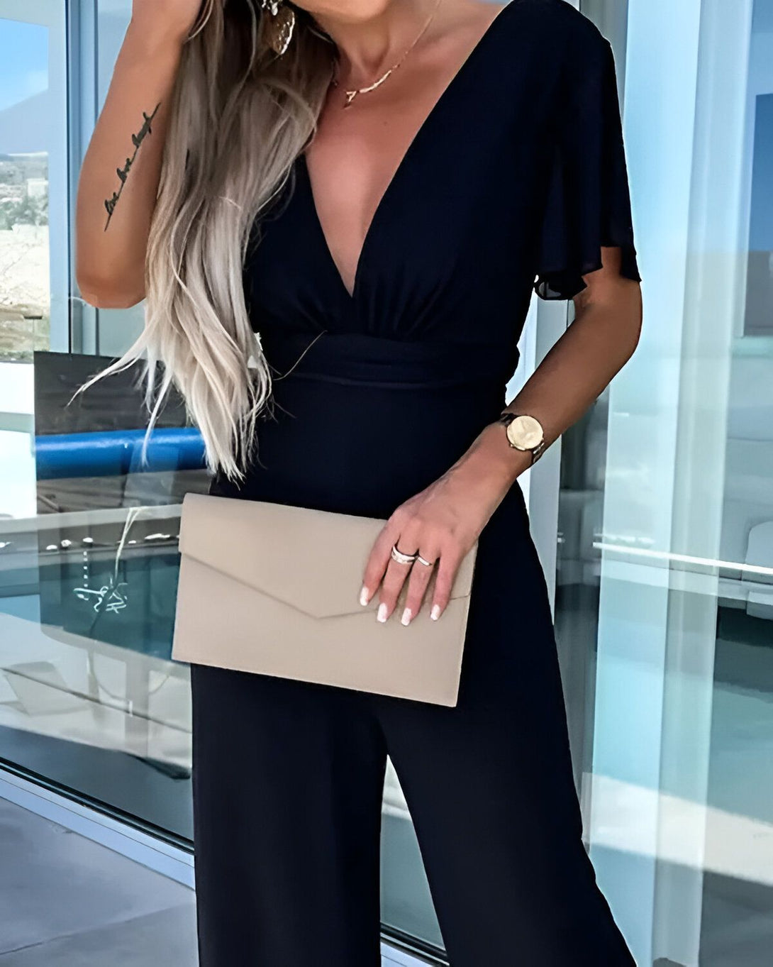 Une femme porte une combinaison noire avec un décolleté en V et des manches courtes. Elle porte également une montre en or et tient une pochette beige à la main.