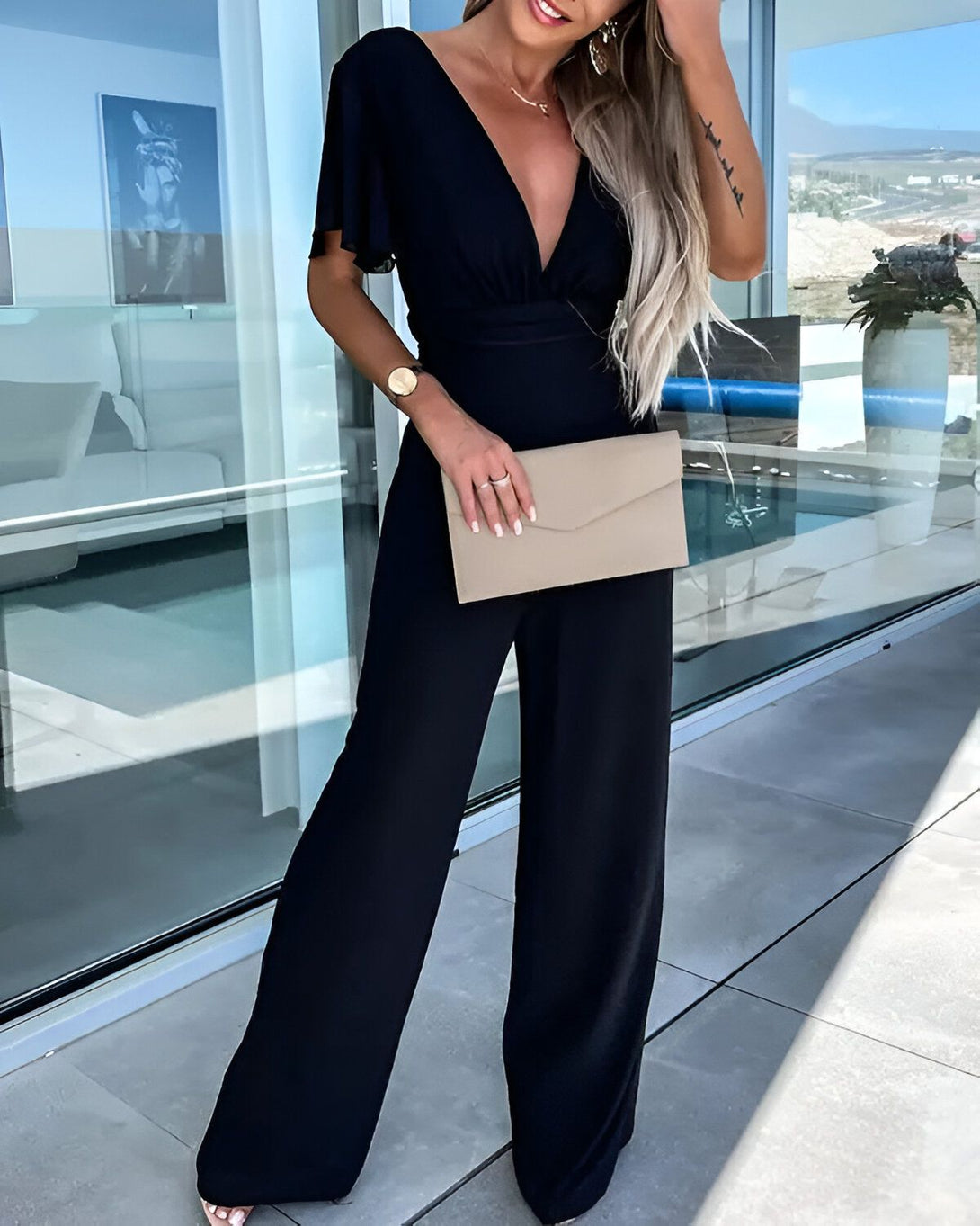 Une femme porte une combinaison noire avec un décolleté en V et des manches courtes. Elle porte également une montre et un collier. Elle tient une pochette beige à la main.