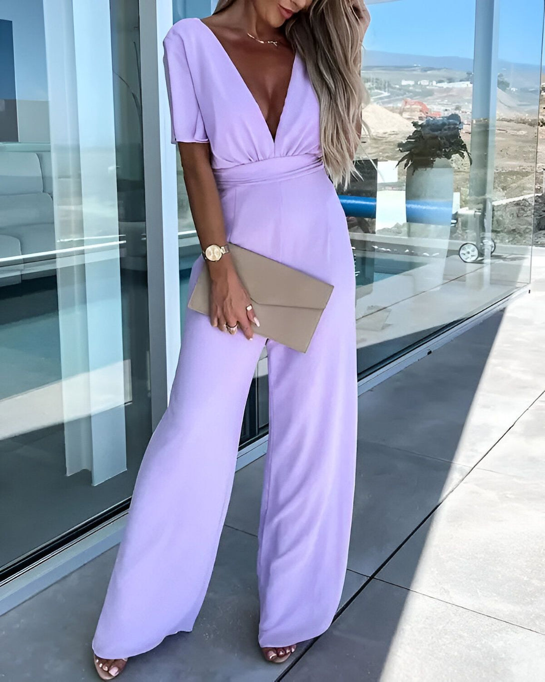 Une femme porte une combinaison pantalon mauve décolleté en V et des sandales à talons transparentes. Elle porte une montre et tient une pochette de couleur beige à la main.