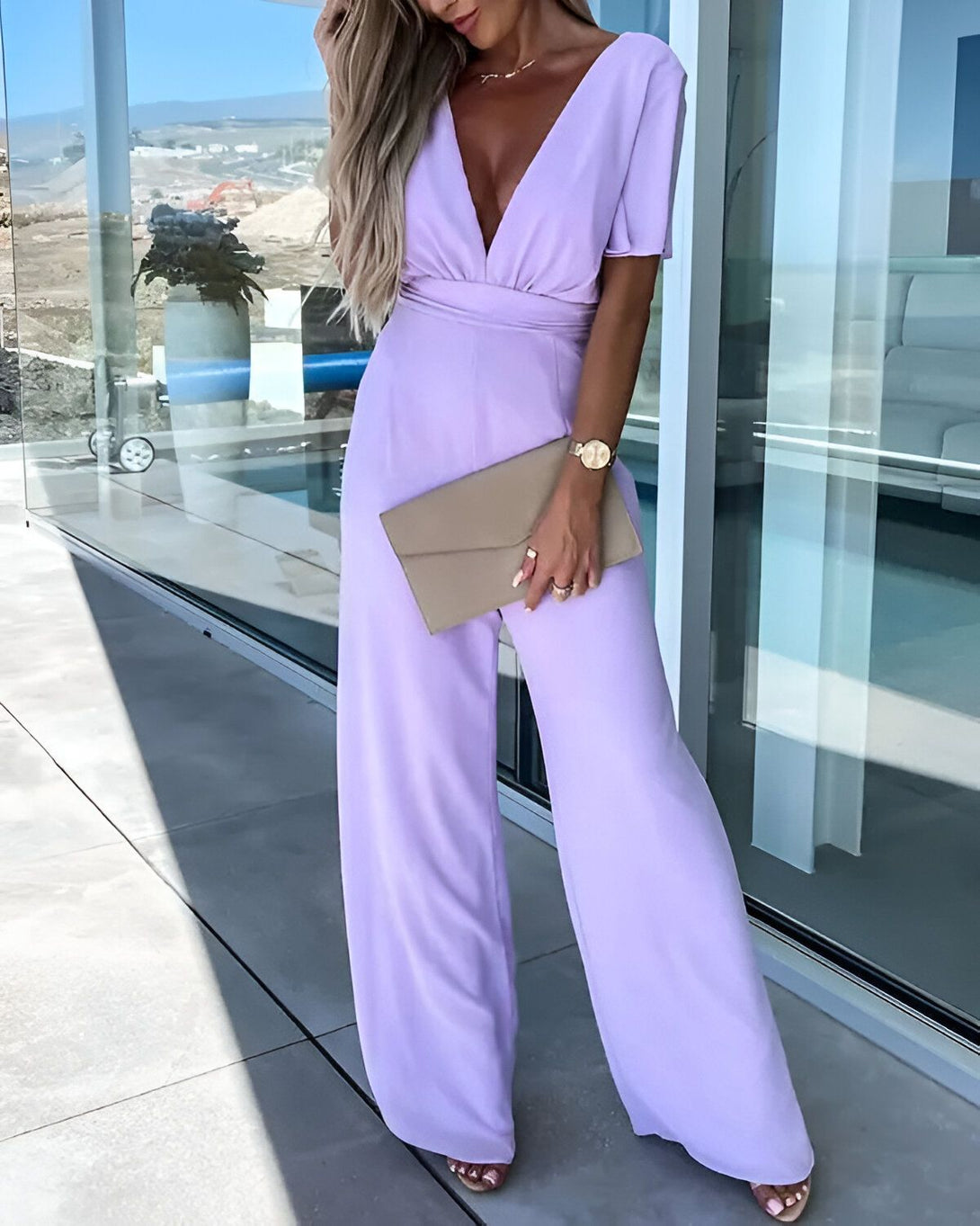 Une femme porte une combinaison pantalon mauve décolleté avec des manches courtes bouffantes. Elle est debout sur une terrasse et porte des sandales à talons hauts couleur chair et un sac à main beige.
