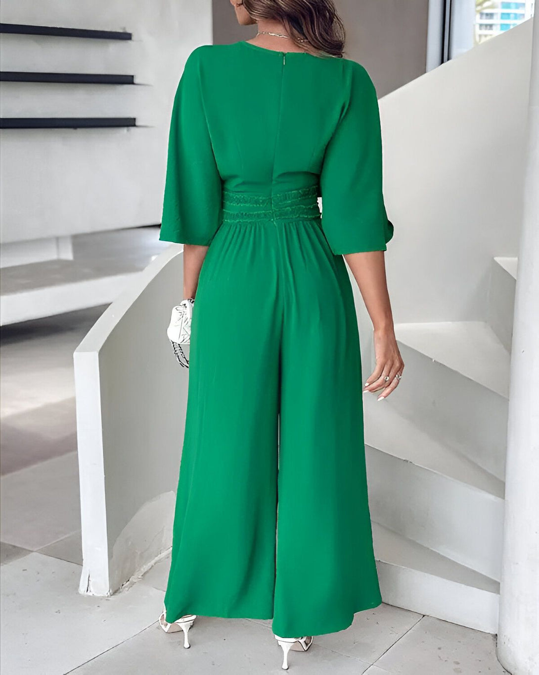 Une femme porte une combinaison pantalon verte. La combinaison a un décolleté en V et des manches bouffantes. Elle est cintrée à la taille et possède des jambes larges. La femme porte des talons blancs et un sac à main blanc.