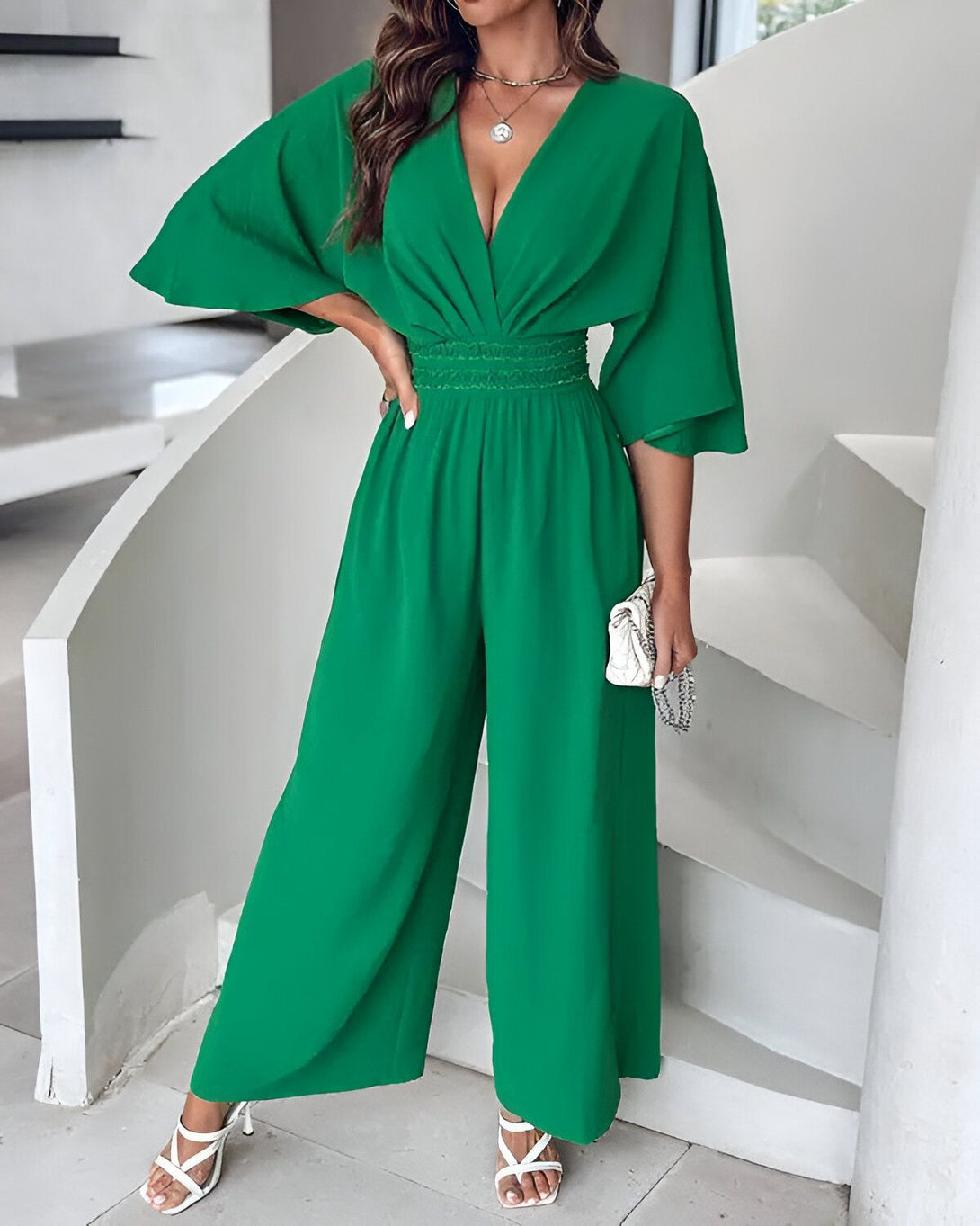 Une femme porte une combinaison-pantalon verte. La combinaison-pantalon est décolletée et possède des manches courtes. Elle est cintrée à la taille et possède des jambes larges. La femme porte des sandales à talons blanches et un collier doré. Elle se tient debout dans un escalier et pose sa main sur sa hanche.