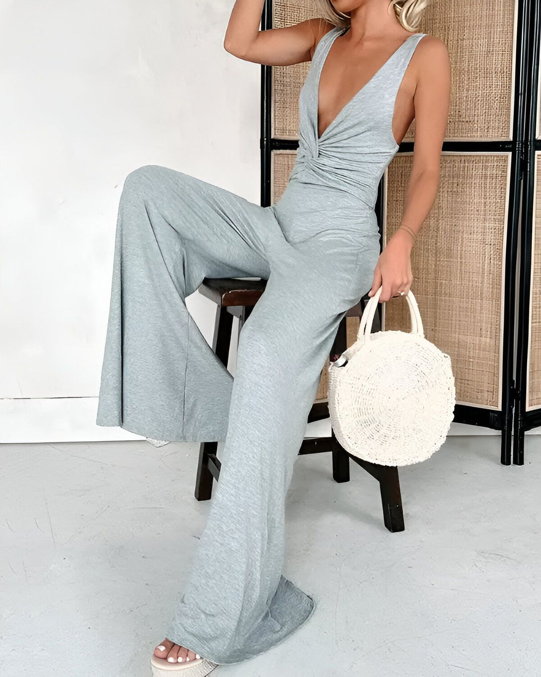 Une femme est assise sur un tabouret, elle porte une combinaison pantalon gris clair et des sandales à talons transparentes. Elle a un sac à main rond en osier à la main droite et porte ses cheveux lâchés.