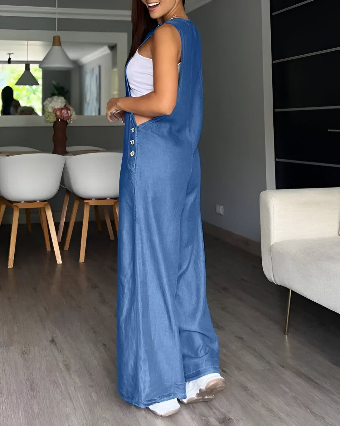 Une femme porte une combinaison-pantalon en jean bleu. Elle est de dos et on peut voir ses cheveux longs et bruns. Elle est dans une pièce avec un sol en parquet et des murs blancs. Il y a une chaise et une table dans la pièce.