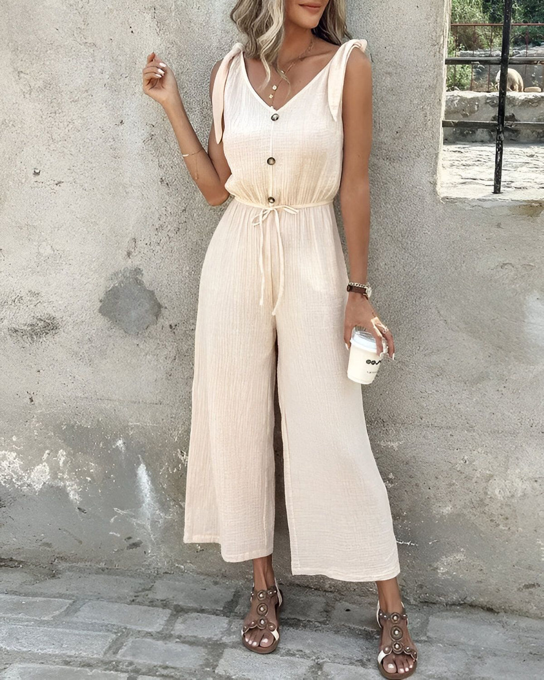 Une femme porte une combinaison-pantalon beige avec des sandales marron clair et tient une tasse à café à la main.
