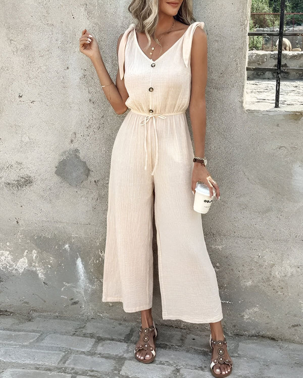 Une femme porte une combinaison-pantalon beige avec des sandales marron clair et tient une tasse à café à la main.