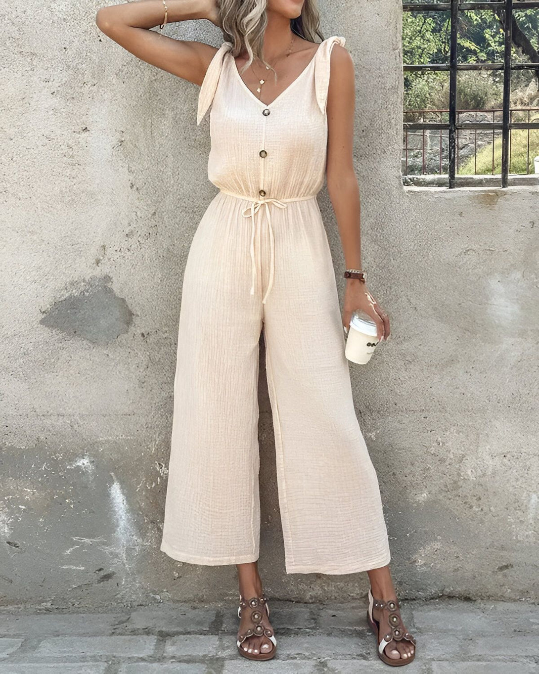 Une femme porte une combinaison-pantalon beige avec des boutons et des bretelles. Elle est pieds nus et porte un café à la main.