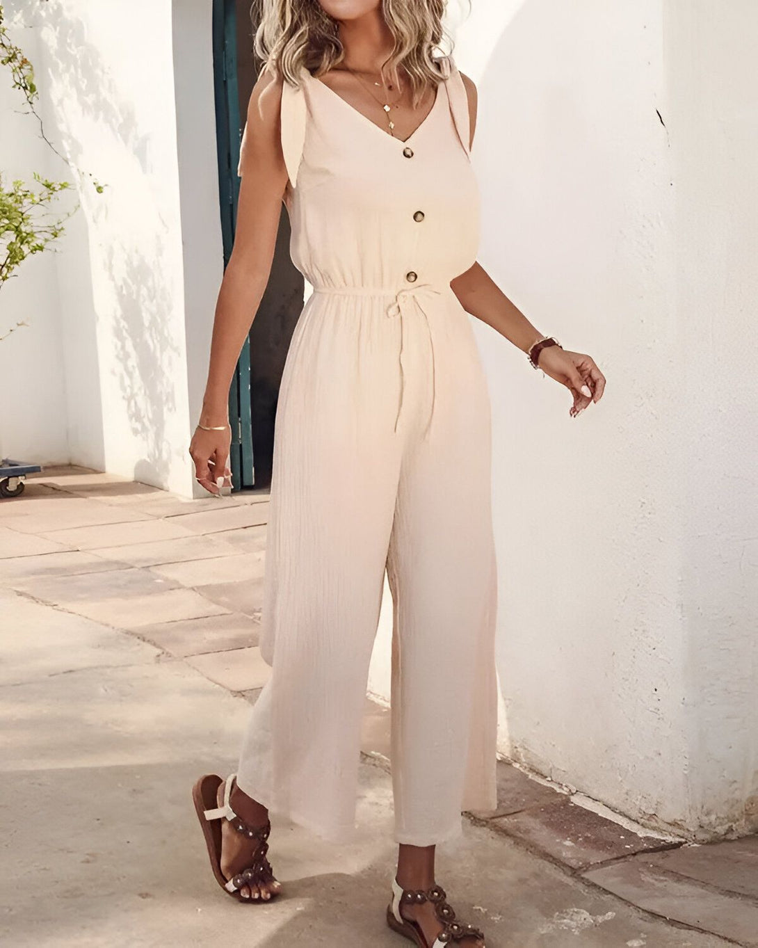 Une femme porte une combinaison-pantalon beige avec des sandales marron. Elle a les cheveux longs et blonds et porte un collier. Elle se tient debout dans une rue et sourit.