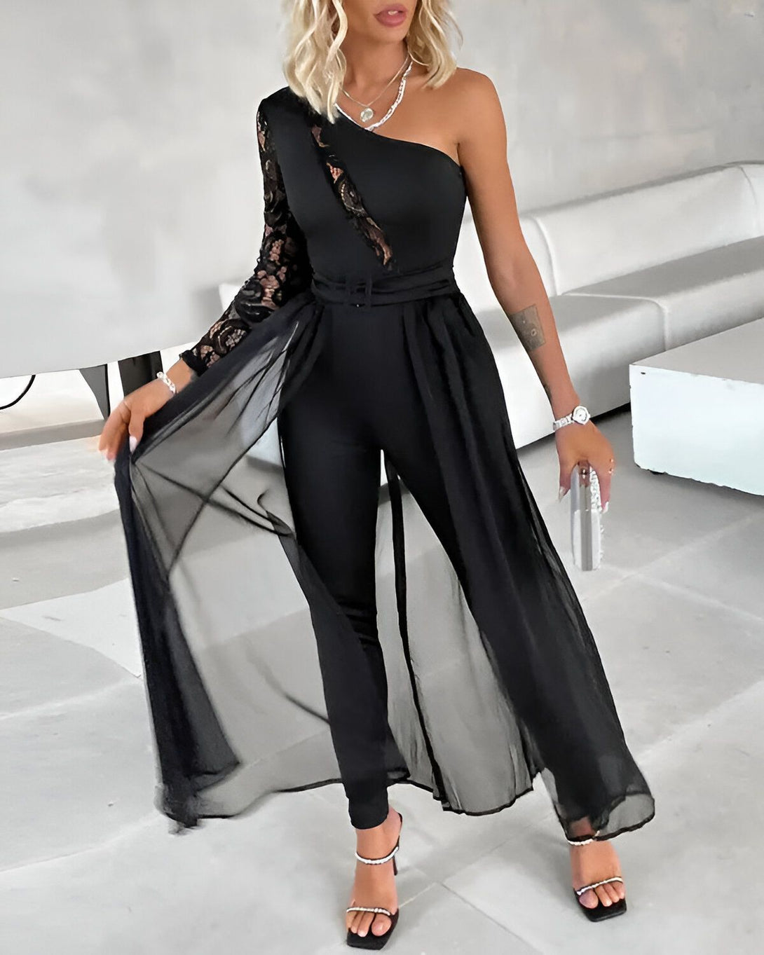 Une femme porte une combinaison noire avec une manche en dentelle et un pantalon large. Elle porte également une ceinture et des talons noirs.