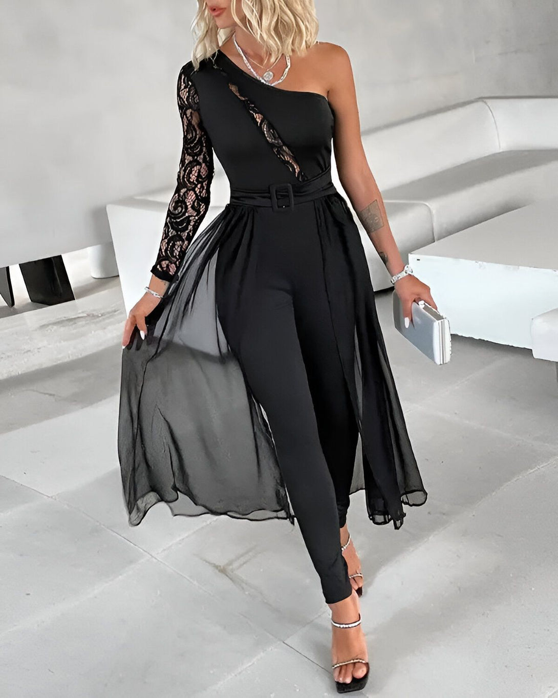 Une femme porte une combinaison noire avec une manche en dentelle et un pantalon large. Elle est également ceinturée et porte des talons hauts noirs.