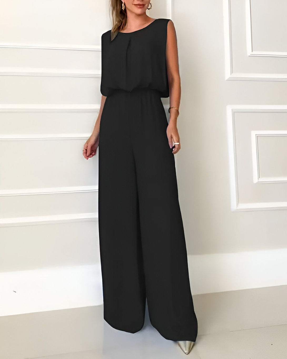 Une femme porte une combinaison-pantalon noire. La combinaison-pantalon est sans manches et a une coupe ample. Elle est ceinturée à la taille. La femme porte des escarpins argentés.