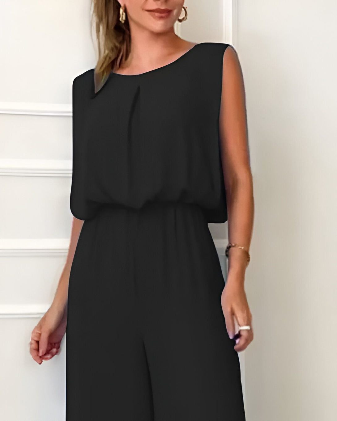 Une femme porte une combinaison noire sans manches. La combinaison est ample et a une taille haute. Elle est également dotée d'un col rond et de poches latérales. La femme est debout, les mains sur les hanches. Elle a les cheveux longs et blonds et porte des boucles d'oreilles.