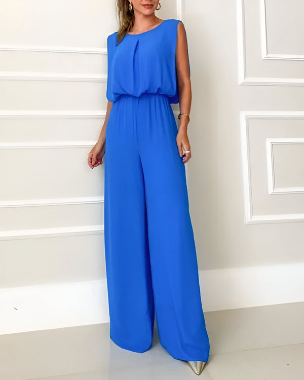 Une femme porte une combinaison-pantalon bleu roi. La combinaison-pantalon est ample et a une coupe décontractée. Elle est dotée d'une encolure ronde et de fines bretelles. La femme porte des talons hauts argentés et un collier doré. Elle a les cheveux longs et blonds et un maquillage léger. Elle se tient debout dans un cadre blanc et regarde l'appareil photo.