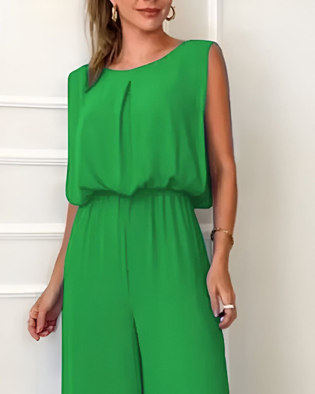 Une femme porte une combinaison pantalon verte. La combinaison est sans manches et a un col rond. Elle est cintrée à la taille et a des jambes larges. La femme est debout et porte des talons hauts blancs. Elle a les cheveux longs et blonds et porte un maquillage léger.