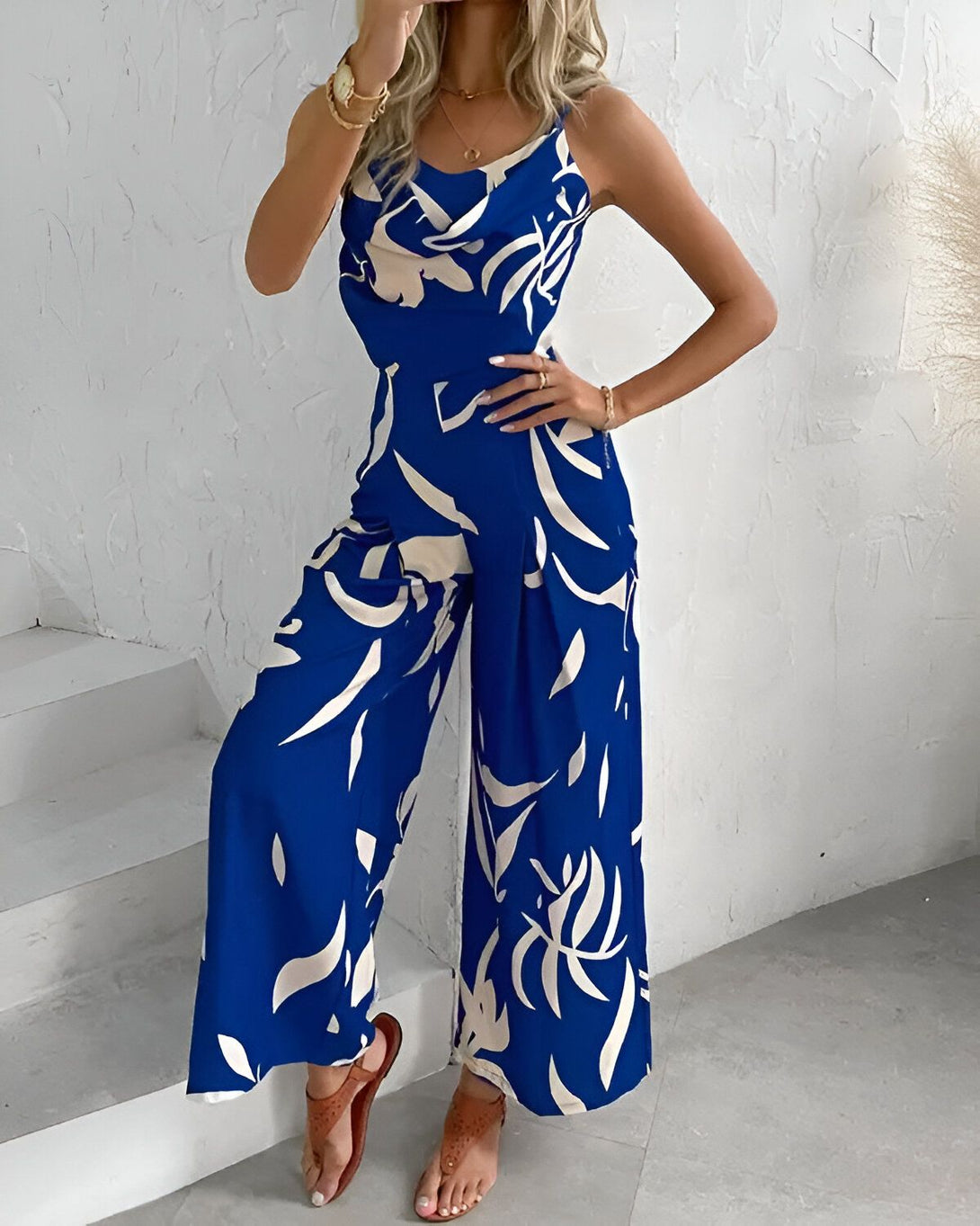 Une femme porte une combinaison pantalon bleue à motifs blancs. La combinaison est décolletée et possède des bretelles fines. Elle est ample et fluide. La femme est blonde et porte des sandales marron. Elle se tient debout sur un fond blanc.