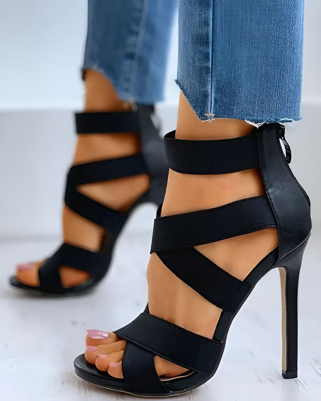 Une femme porte des sandales à talons hauts noires. Les sandales sont faites de lanières de tissu noir qui s'entrelacent sur le pied. Le talon est fin et mesure environ 10 centimètres. La femme porte un jean bleu troué au niveau des genoux.