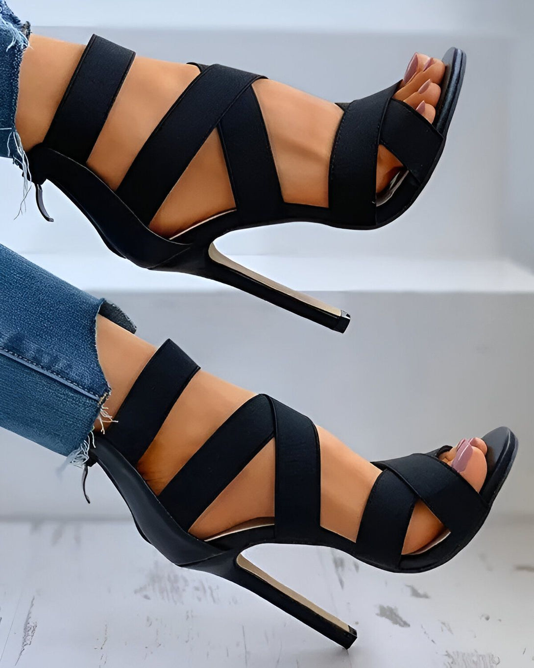 Une femme porte des sandales à talons hauts noires. Les sandales sont faites de lanières de tissu qui s'entrelacent sur le pied. Le talon est fin et mesure environ 10 centimètres. La femme porte un jean bleu déchiré et les ongles des pieds peints en rose.