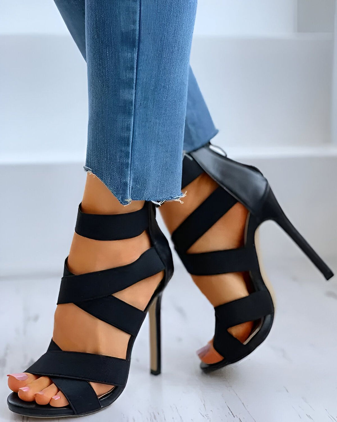 Une femme porte des sandales à talons hauts noires. Les sandales sont faites de lanières de tissu noir qui s'enroulent autour de la cheville et du pied. La femme porte un jean bleu troué au niveau des genoux.