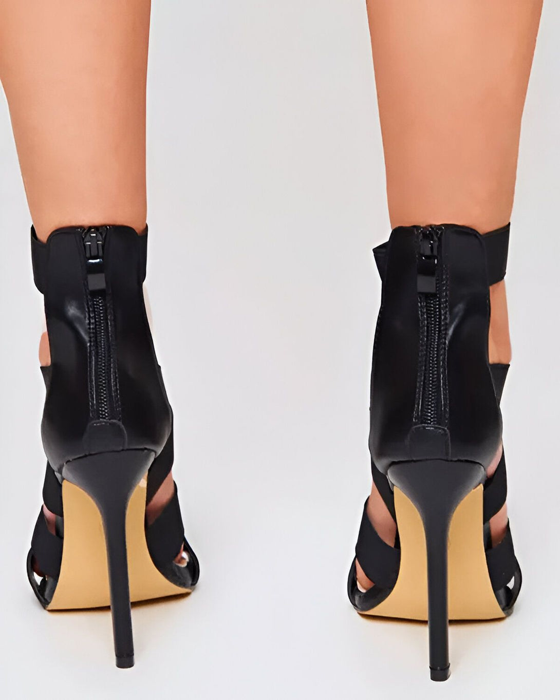 Une image de dos de deux jambes de femme portant des sandales noires à talons hauts. Les sandales ont des brides fines et un bout ouvert. Les talons sont fins et dorés.