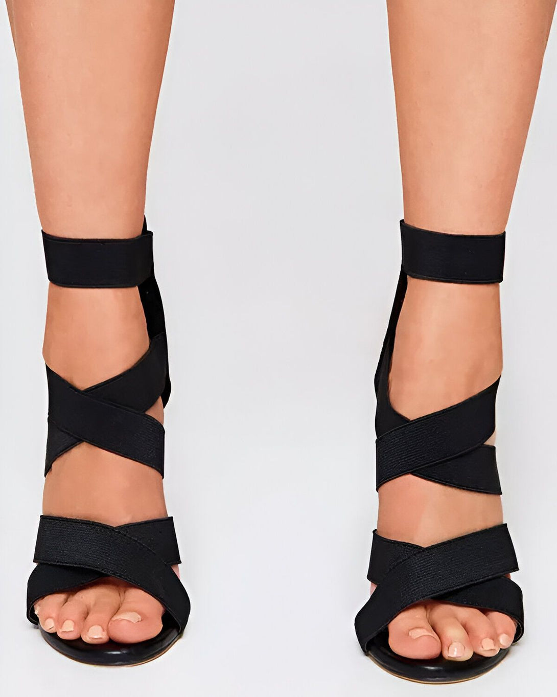 Une femme porte des sandales noires à talons hauts. Les sandales sont faites de lanières de tissu élastique qui s'enroulent autour de la cheville et du pied. Les talons sont fins et hauts d'environ 10 centimètres. La femme a les ongles des pieds peints en rose.