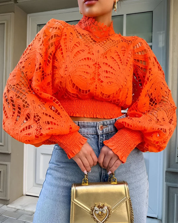 Une femme porte un chemisier orange en dentelle avec un col montant et des manches bouffantes. Elle porte également un jean bleu et un sac à main doré.