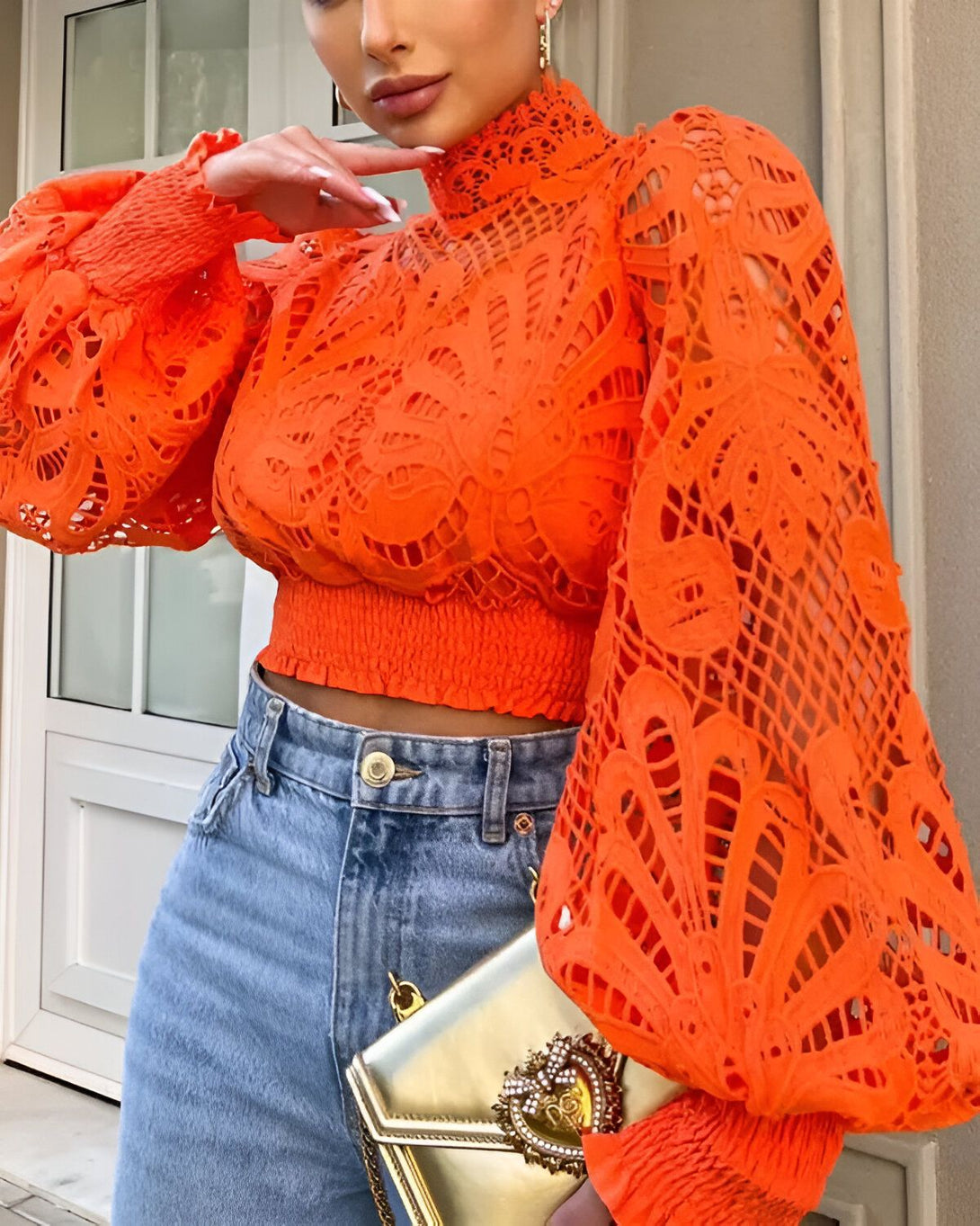 Une femme porte un chemisier orange à manches bouffantes et un jean bleu. Elle a une main sur la hanche et l'autre tenant un sac à main doré.