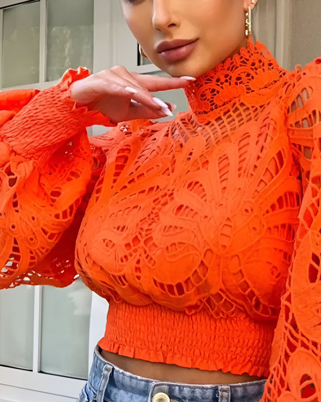 Une femme porte un chemisier orange en dentelle avec un col montant et des manches bouffantes. Elle a les cheveux noirs et les lèvres nues. Elle porte également un jean bleu.