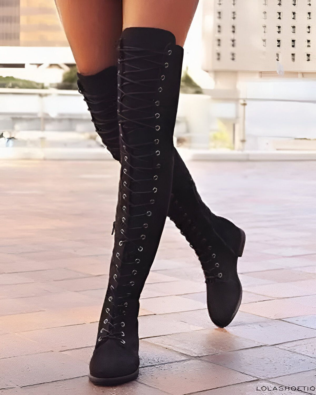 Une femme porte des bottes noires à lacets. Les bottes sont hautes et montent jusqu'au-dessus du genou. La femme est debout sur un sol en pierre.
