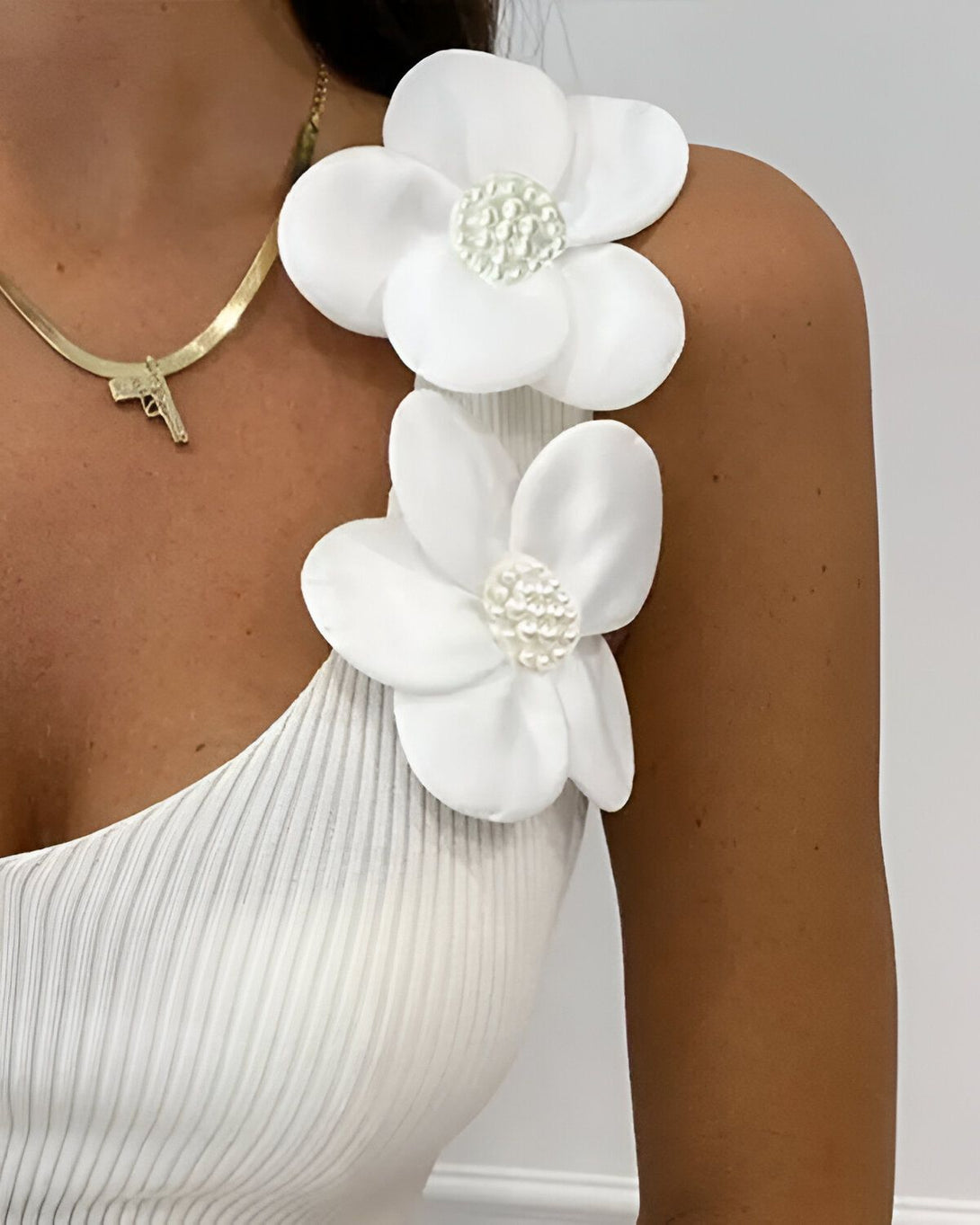 Une femme porte un haut blanc à bretelles avec des fleurs blanches en tissu sur l'épaule. Elle porte également un collier doré avec un pendentif en forme de pistolet.