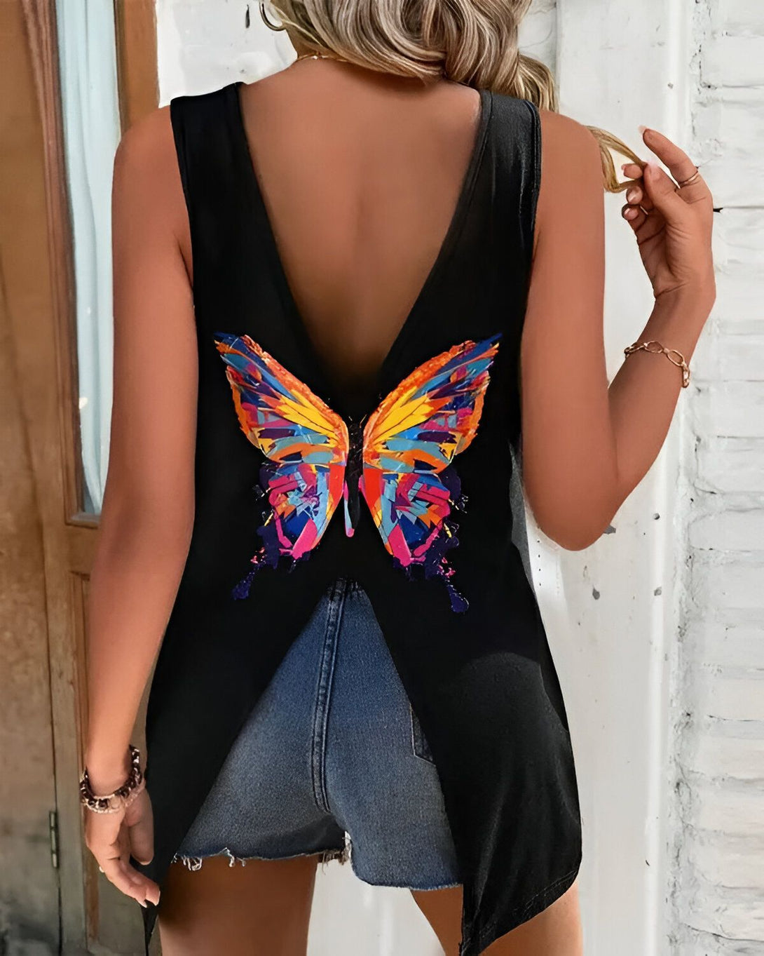 Une femme porte un débardeur noir avec un papillon coloré dans le dos. Le papillon est peint en orange, jaune, bleu et vert. La femme est vue de dos et porte un short en jean bleu.