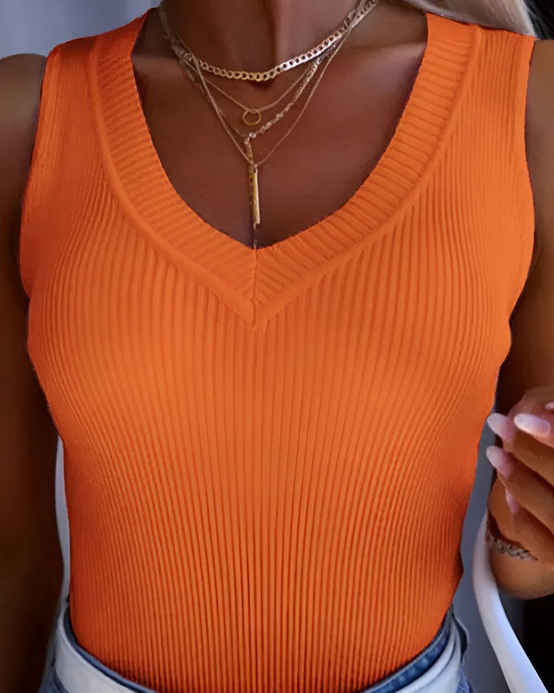 Une femme porte un débardeur orange côtelé avec un col en V profond. Elle porte également plusieurs colliers et un bracelet.