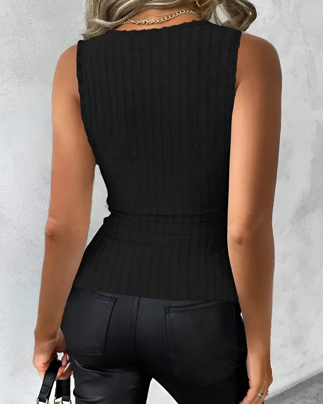 Une femme porte un débardeur côtelé noir. Le débardeur est cintré à la taille et possède un col montant. La femme porte également un pantalon en cuir noir et un collier doré.