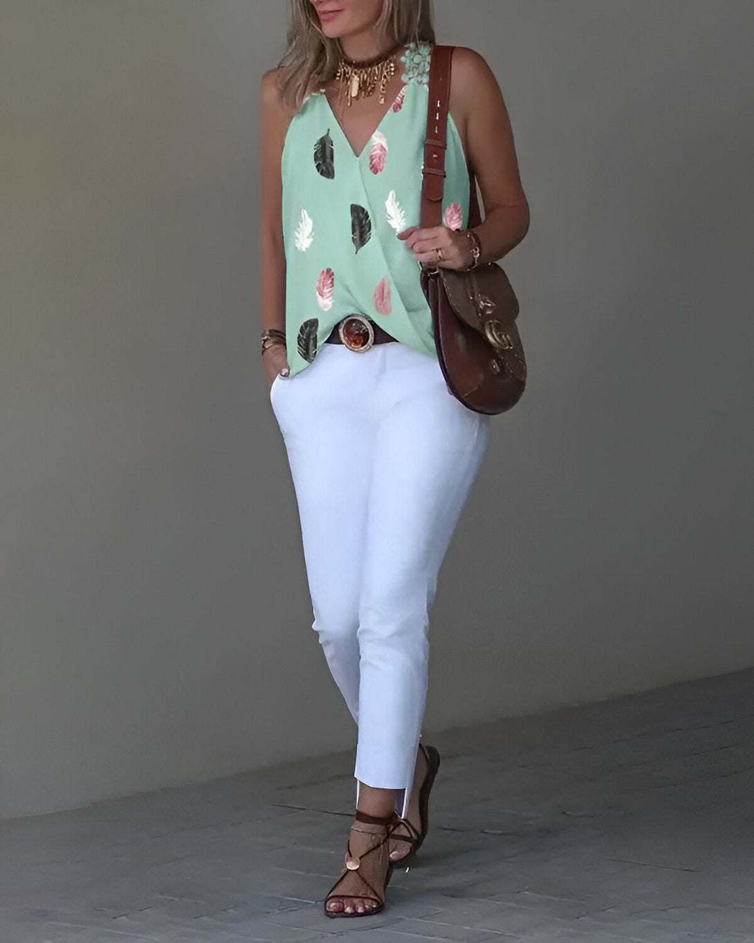 Une femme porte un débardeur vert à motifs de feuilles tropicales et un pantalon blanc. Elle a un sac à main marron et des sandales marron.