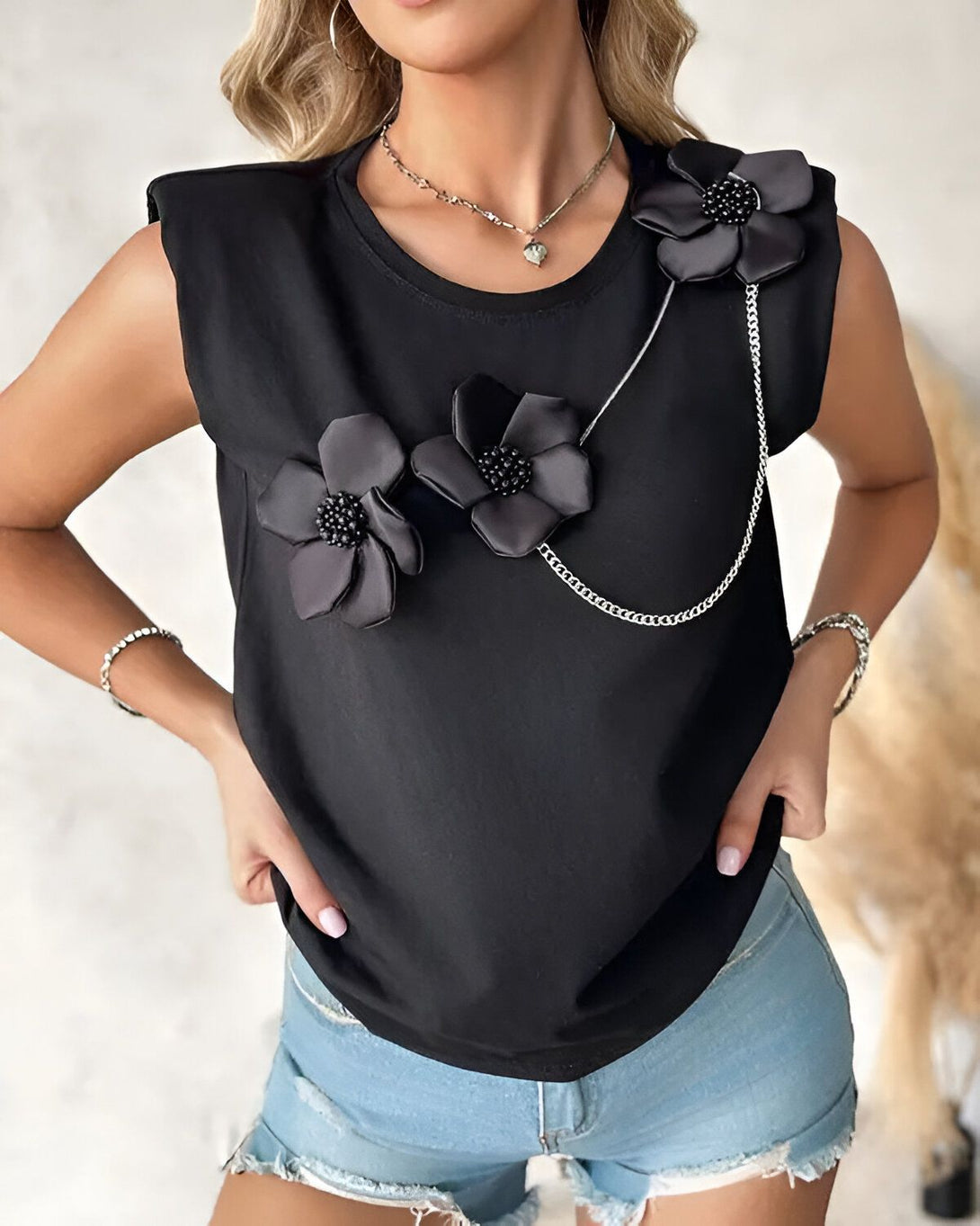 Une femme porte un t-shirt noir sans manches avec des fleurs en satin noir et une chaîne sur l'épaule. Elle porte également un short en jean bleu clair et des sandales noires.