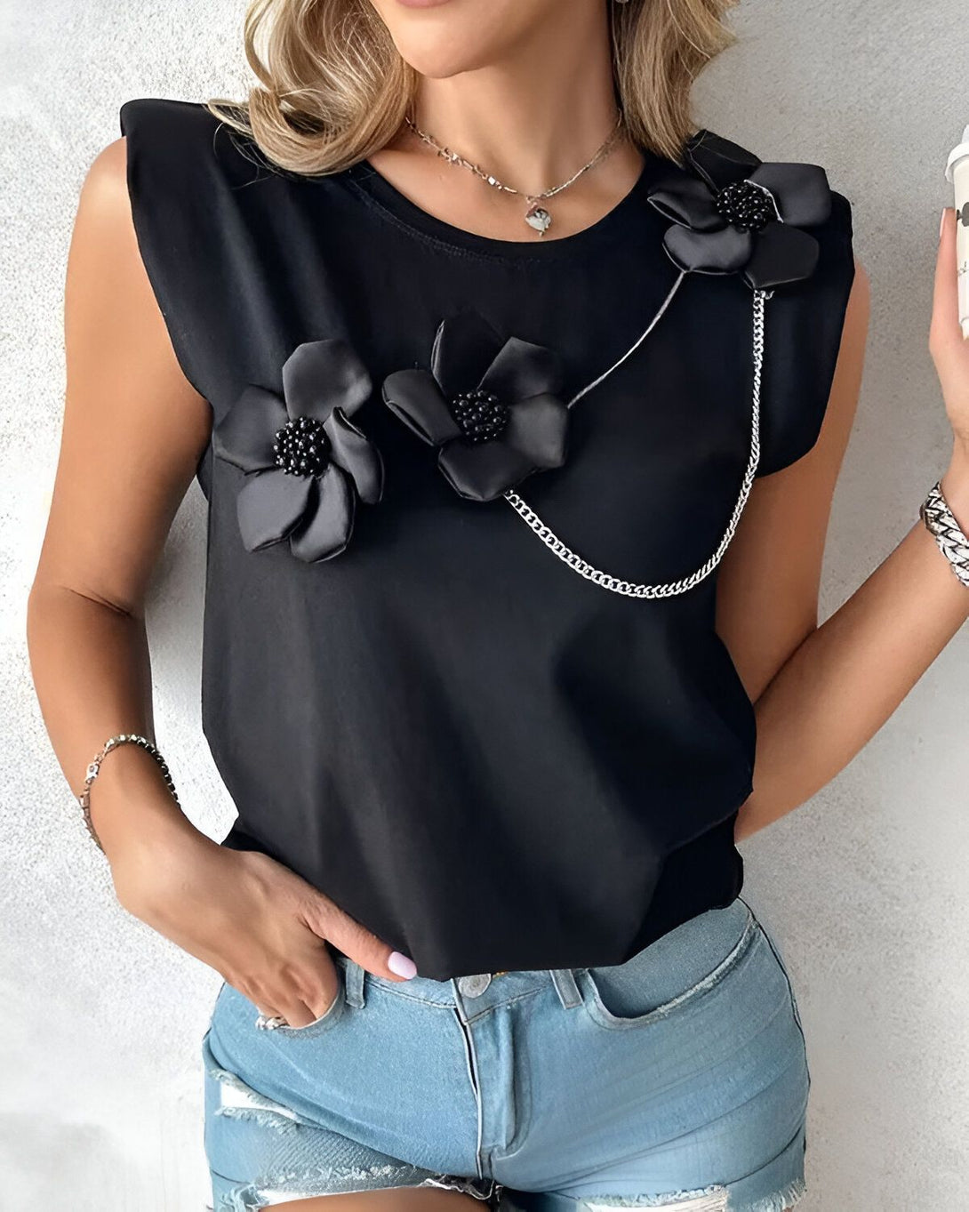 Une jeune femme porte un t-shirt noir sans manches avec des fleurs en tissu noir et des chaînes. Elle porte également un jean bleu déchiré et un collier avec un pendentif en forme de cœur.
