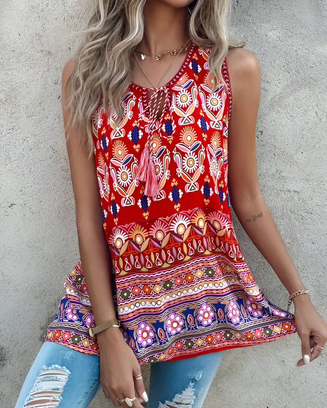 Une jeune femme porte un débardeur rouge sans manches à motifs ethniques. Le débardeur est évasé et possède un lien à nouer au niveau du cou. La femme porte également un jean bleu troué et des sandales marron.