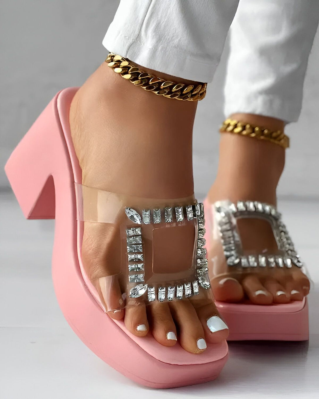 Une femme porte des mules à talons roses avec des brides transparentes et des strass. Elle porte également un jean blanc et un bracelet de cheville doré.