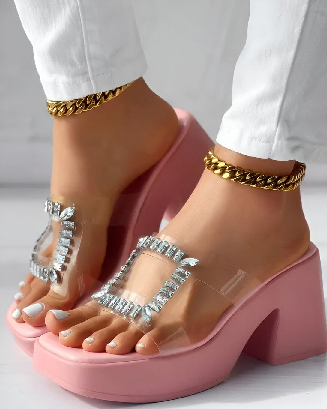 Une femme porte des sandales à talons roses transparentes avec des strass. Elle porte également un jean blanc et des bracelets de cheville en chaîne dorée.
