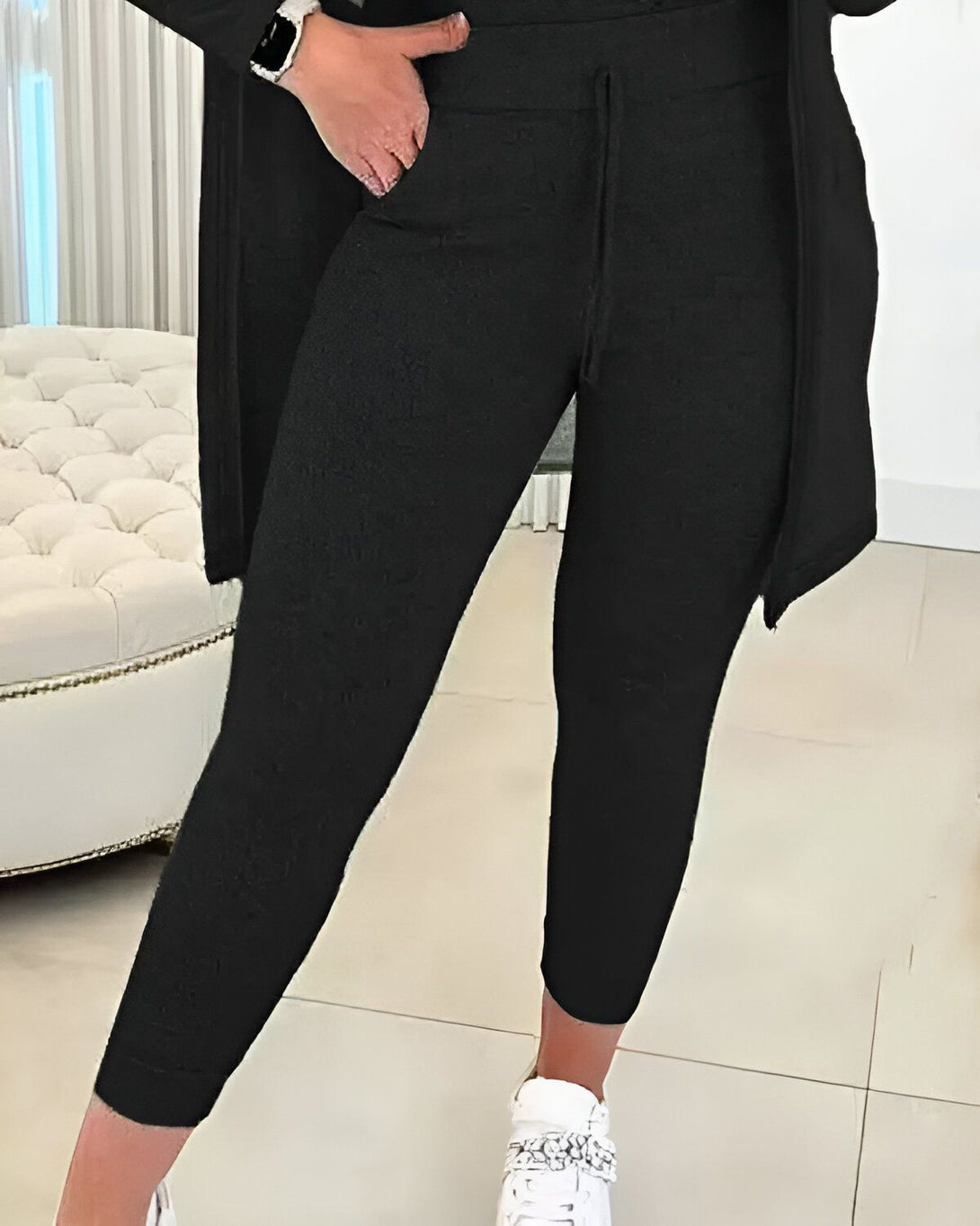 Une femme porte un pantalon de jogging noir et une veste ouverte noire. Elle est debout dans une pièce blanche et porte des baskets blanches.