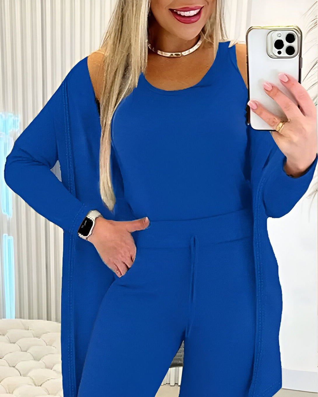 Une femme porte un ensemble trois pièces bleu composé d'un débardeur, d'un pantalon et d'un cardigan. Elle a une montre au poignet gauche et tient un téléphone portable dans sa main droite.