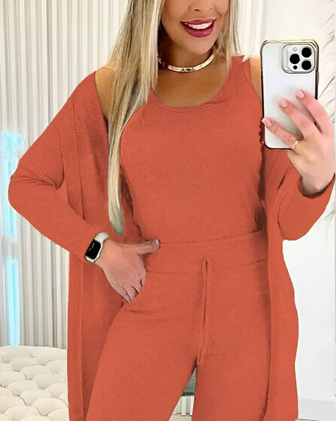 Une femme porte un ensemble de jogging orange composé d'un débardeur, d'un pantalon et d'un cardigan. Elle est blonde, souriante et tient son téléphone portable à la main.