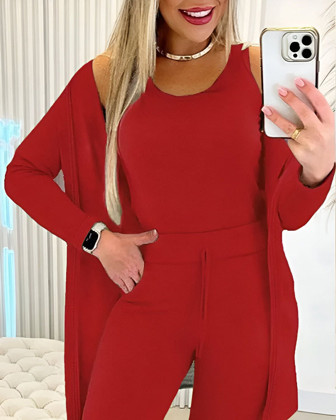 Une femme porte un ensemble trois pièces rouge composé d'un débardeur, d'un pantalon et d'un cardigan. Elle a une montre connectée au poignet gauche et tient un téléphone portable dans sa main droite.